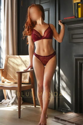 Slim English redhead in red panties and bra standing by a fireplace